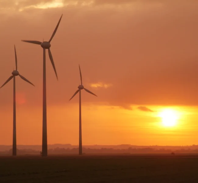 Three wind turbines silhouetted against an orange sunset sky over a flat, open landscape.