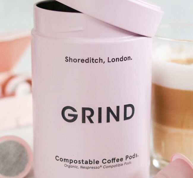Pink Grind tin labeled “Compostable Coffee Pods” with lid open, surrounded by coffee pods and a glass of coffee.