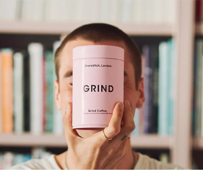 Person holding a pink GRIND coffee canister in front of their face, with a blurred bookshelf in the background.