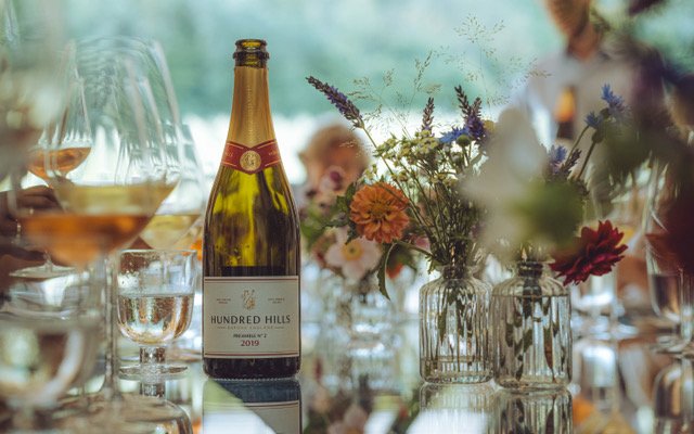 Bottle of Hundred Hills wine on table with glassware and wildflower vases at an outdoor celebration.