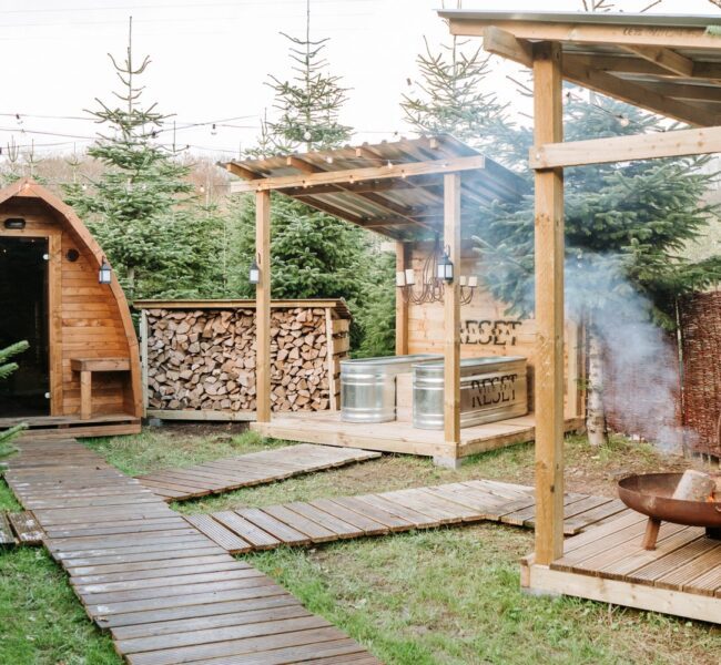 Outdoor glamping area with a wooden pod cabin, fire pit, hot tub, seating deck and string lights among evergreen trees.