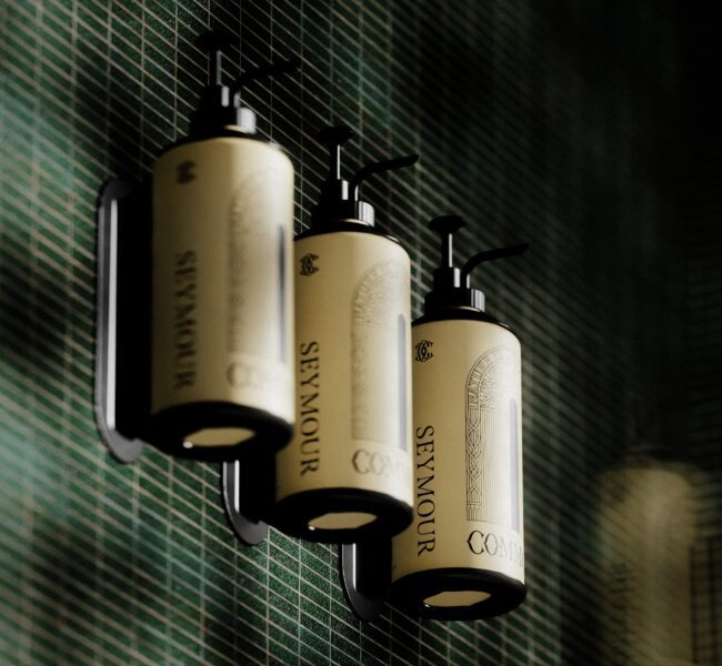 Three cream Seymour toiletry bottles with black pumps mounted on a dark green tiled wall in a black wall bracket.